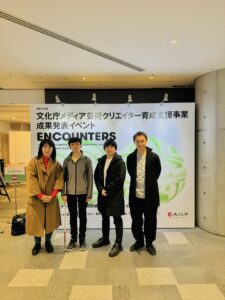 「文化庁メディア芸術クリエイター育成支援事業 成果発表イベント『ENCOUNTERS』」の会場前で撮影された写真。背景にはイベントのパネルがあり、4人のメンバーがその前に並んでいる。全体的にフォーマルで落ち着いた雰囲気で、4人は穏やかに笑っている。