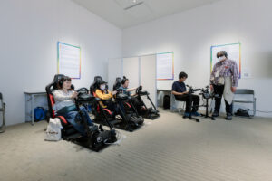 展示風景。専用のドライビングシートに腰掛けた参加者たちは、ヘッドホンを装着し、ハンドルとペダルを操作している。その横では、電子ドラムをプレイする参加者の姿も見られ、視覚に依存しない多様なゲーム体験がこの空間で並列的に展開されていることがわかる。