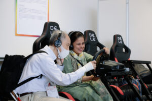 視覚に障害のある外国人の女性がのレーシングゲームを体験している様子。女性はヘッドホンを装着し、笑顔を浮かべながらハンドルを操作しており、隣に座る支援者が優しく寄り添っている。